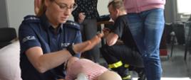 Szkolenie z pierwszej pomocy. Świetlica Komendy Powiatowej Policji w Rawiczu. Na pierwszym planie umundurowana funkcjonariuszka policji, która klęcząc na prawym kolanie, na lewym trzyma manekina "niemowlaka" - wykonując czynności ratujące życie po uprzednim zakrztuszeniu się dziecka. 