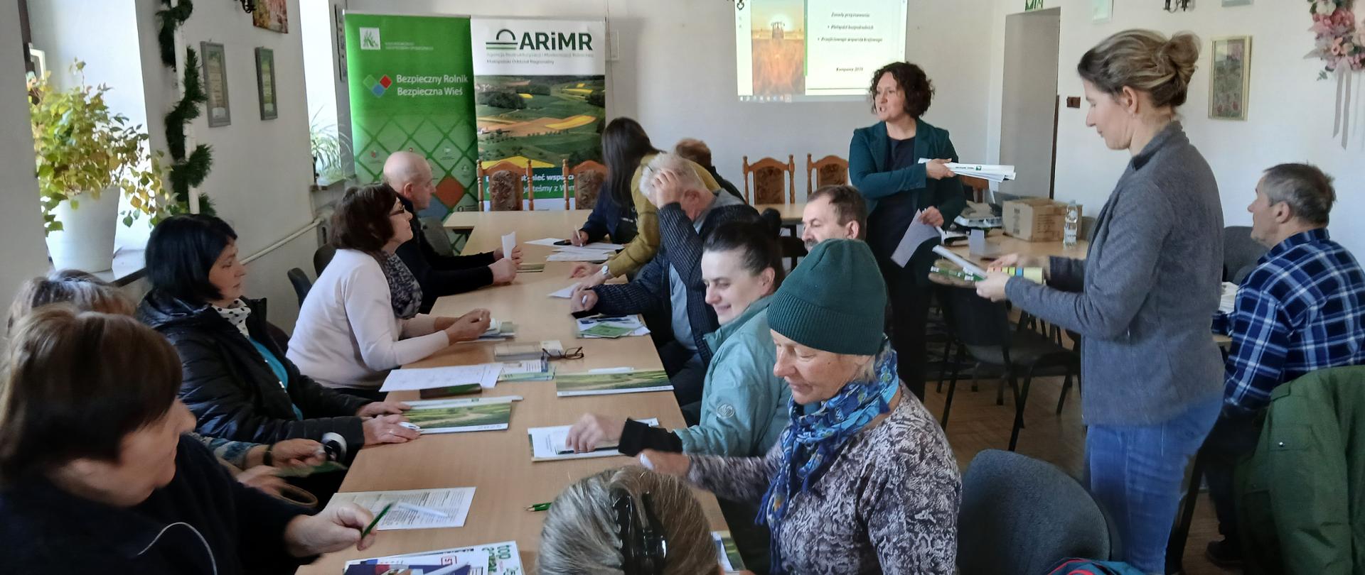 Widok na salę szkoleniową, grupa osób siedząca przy stole, na stole dokumenty, ulotki