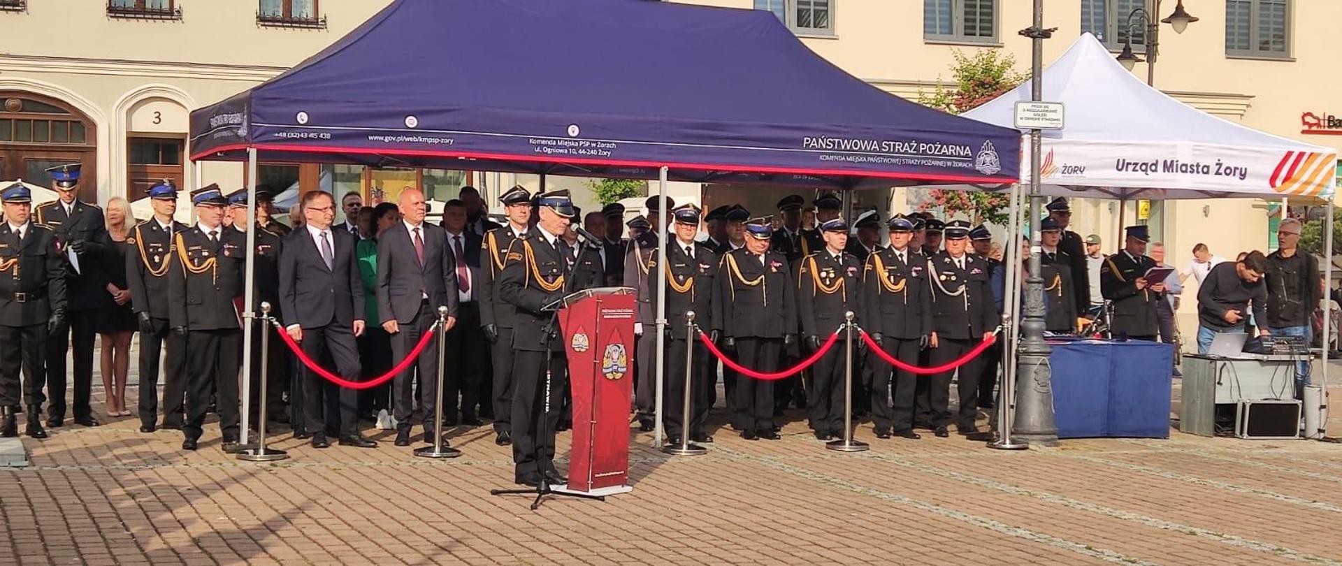 Strażąk w mundurze galowym stoi przed mównicą. Za nim pod zadaszeniem w kolorze granatowym stoją goście. W tle widać budynki.