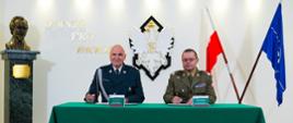 Szef ITD i Rektor-Komendant WAT siedzią przy zielonym stole i podpisują dokumenty. W tle rzeźba z patronem uczelni gen. Jarosławem Dąbrowskim, biały orzeł, godło Polski, napis „Omnia Pro Patria” oraz flagi Polski i NATO.