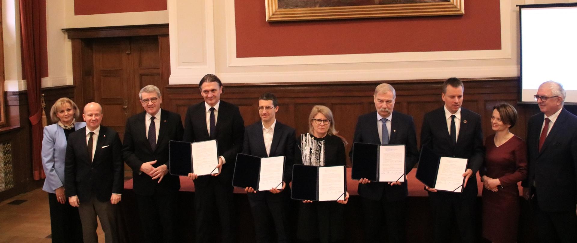 10 osób stoi przed stołem, pokazują trzymane w rękach dokumenty, za nimi na czerwonej ścianie obraz.