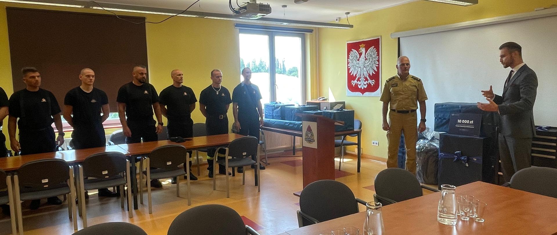 W sali wykładowej Komendy Miejskiej Państwowej Straży Pożarnej w Wałbrzychu. Po lewej stronie przy mównicy stoi w mundurze piaskowym Komendant Miejski PSP w Wałbrzychu, obok w ciemnym garniturze Prezes Wałbrzyskiej Specjalnej Strefy Ekonomicznej. Po lewej stronie przy stole stoją strażacy