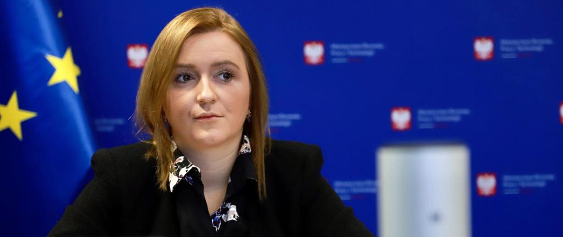 Wiceminister Olga Semeniuk podczas konferencji Kompetencje jutra - Lublin inspiruje przyszłość”. Siedzi przy stole, za wiceminister widać baner ministerstwa rozwoju.