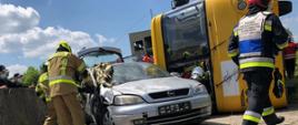 Wypadek samochodowy na pierwszym planie srebrny samochód osobowy z którego strażacy ewakuują osobę poszkodowaną. Za samochodem na boku leży autokar do środka wchodzą strażacy.