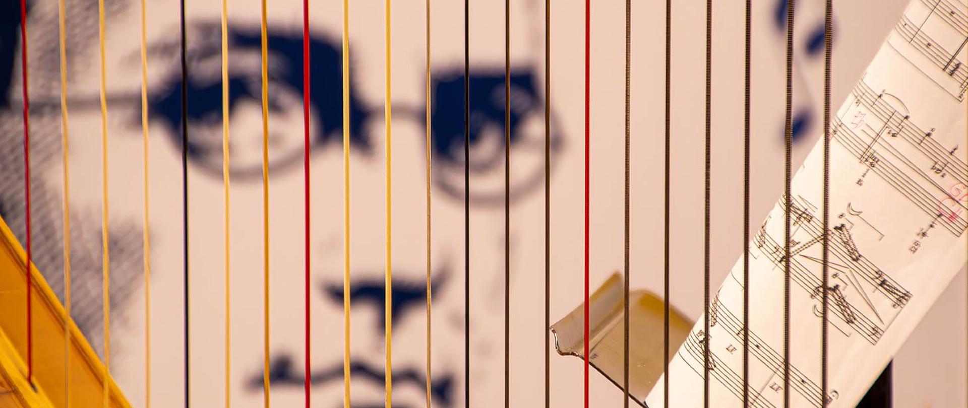 Na zdjęciu struny harfy, za nimi z prawej strony pulpit z nutami i dalej z tyłu rozmyty rysunek Moniuszki z baneru szkolnego.