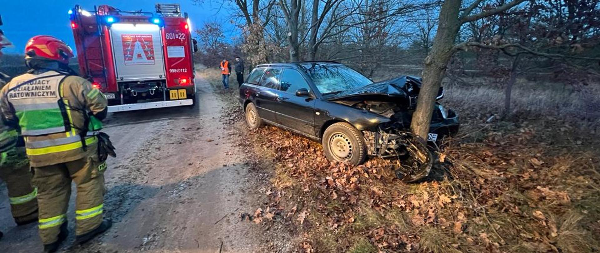 Wrak pojazdu na poboczu z widocznymi uszkodzeniami przedniej części pojazdu, w tyle samochód strażacki obok wraku strażacy