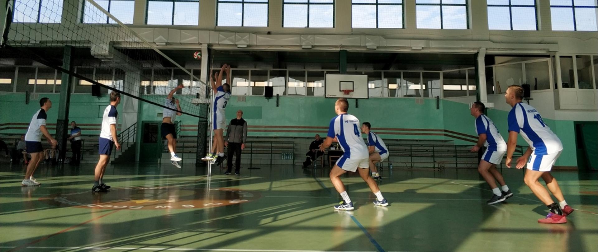 Zawodnicy w biało-niebieskich stronach podczas gry w siatkówkę w hali gimnastycznej, na zielonej nawierzchni. Przy siatce kilku wyskakuje do zablokowania piłki, zawodnik drużyny przeciwnej atakuje.