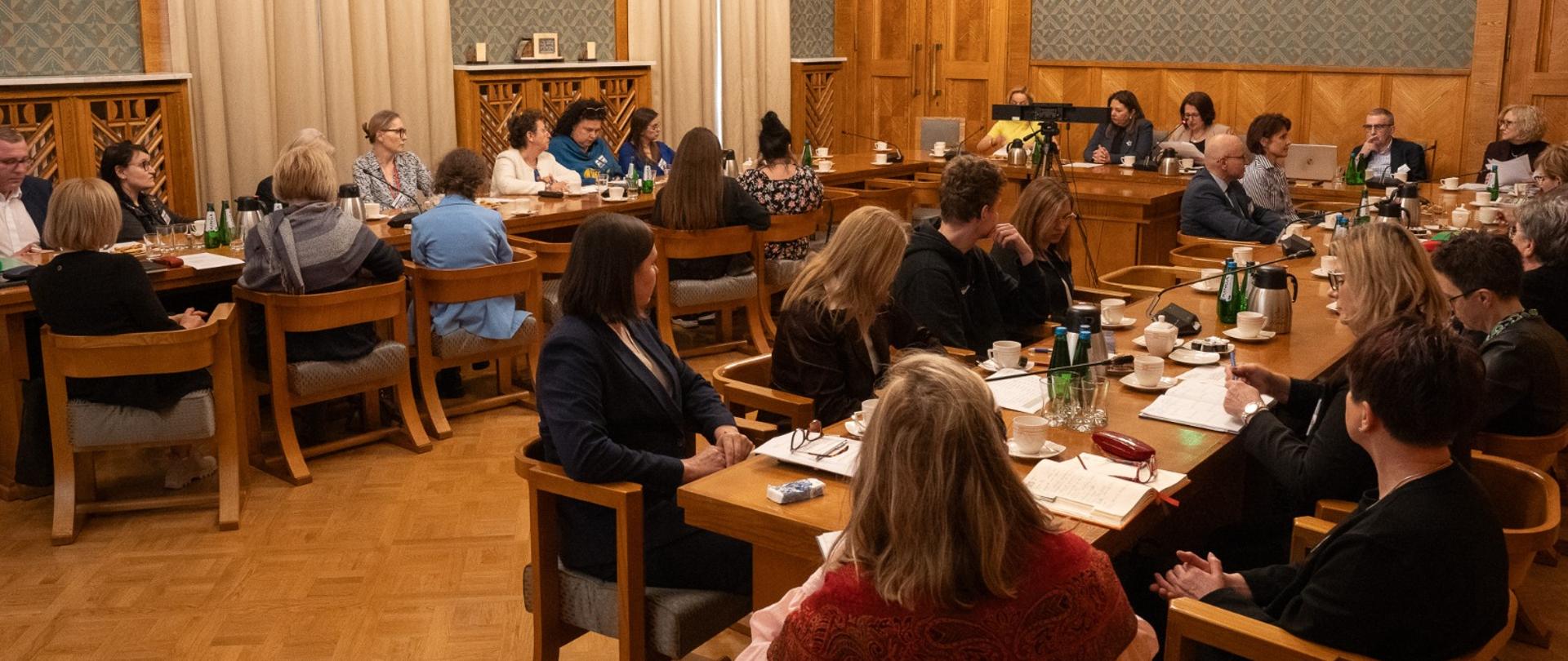 Uczestnicy spotkania siedzą przy dwóch stołach po lewej i po prawej stronie sali. Prostopadle do nich, na wprost od wejścia stoi stół, przy którym siedzi m.in. minister Izabela Ziętka i prowadzi spotkanie. Uczestnicy przysłuchują się wypowiedzi minister i kierują na nią spojrzenie. Na stołach leżą kartki, zeszyty, dokumenty, stoją butelki z wodą i filiżanki. 