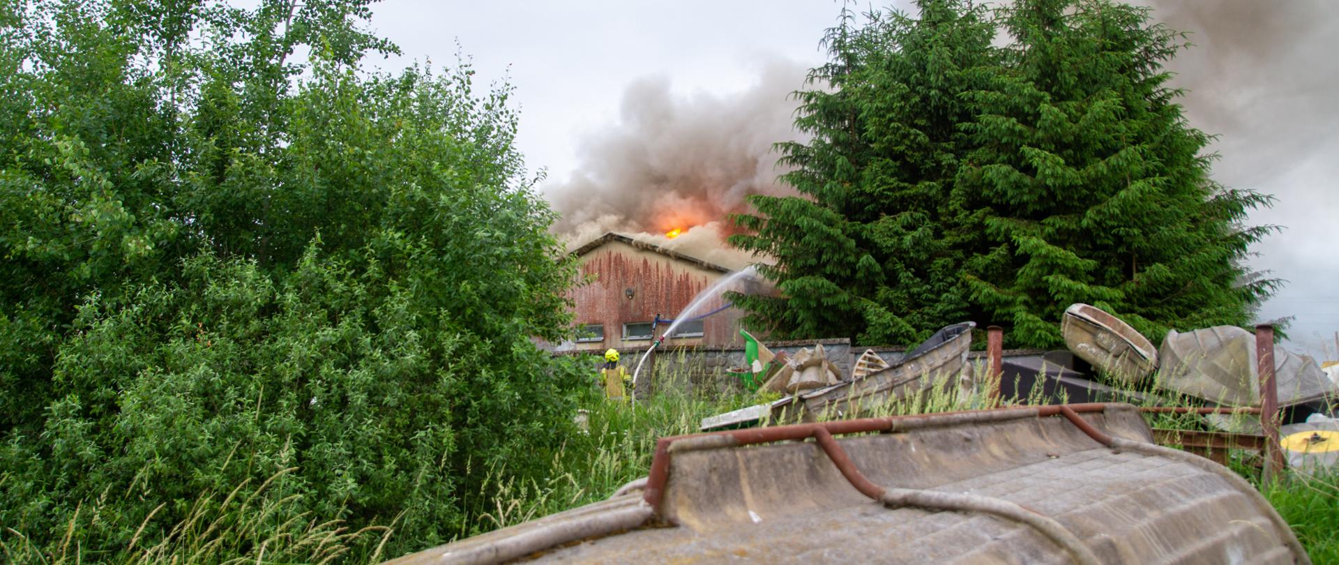 Na pierwszym panie zarys łodzi, odwróconej do góry dnem. W oddali zdjęcia w zaroślach jeden strażak podaję wodę przez ogrodzenie na widoczny dalej zarys budynku. Widoczne zarysy ognia i kłęby dymu. 
