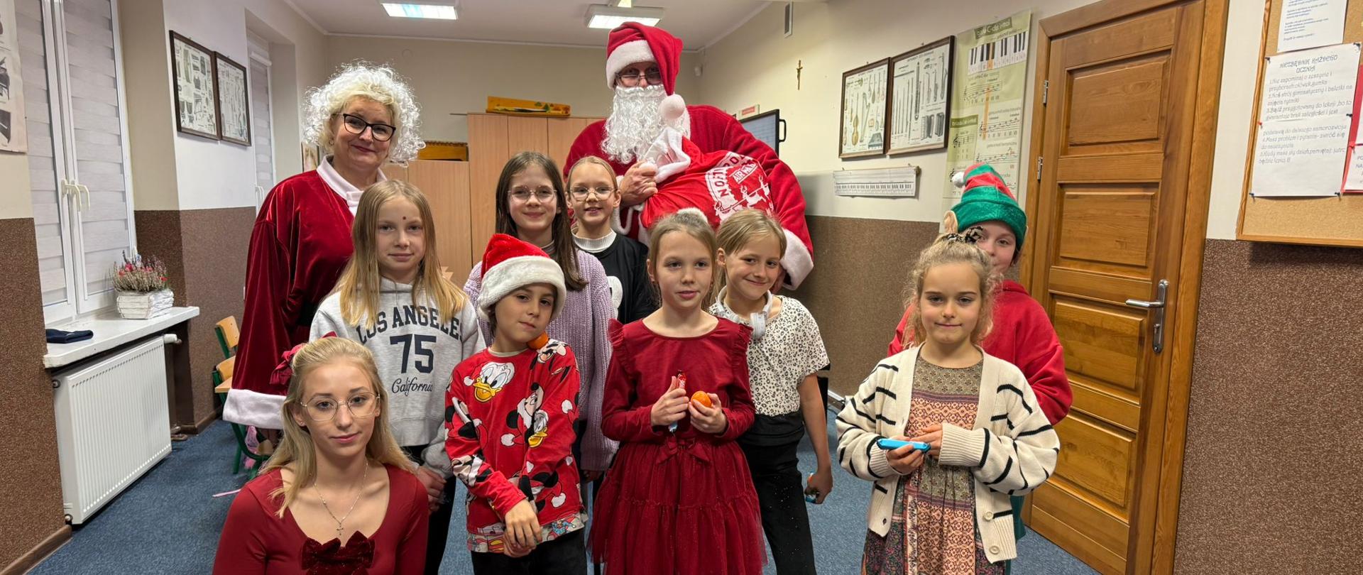 W szkolnej sali muzycznej Święty Mikołaj w czerwonym stroju obok niego dwie kobiety blondynki w okularach ubrane na czerwono oraz grupa uczniów W tle widać szkolne wyposażenie.
