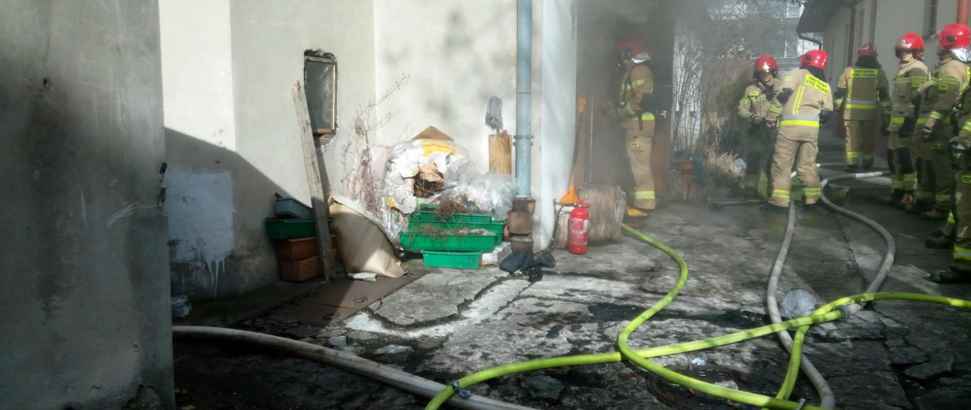 Zdjęcie przedstawia podwórze wewnętrzne zabudowań przy ulicy Leonarda w Kielcach. Widać silne zadymienie oraz rozciągnięte węże gaśnicze, a głębiej przemieszcza się grupa pięciu strażaków.