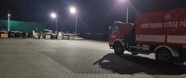 Samochody ciężarowe straży pożarnej oczekują na rozładunek, niedaleko znajduje się wypakowany sprzęt pożarniczy.
