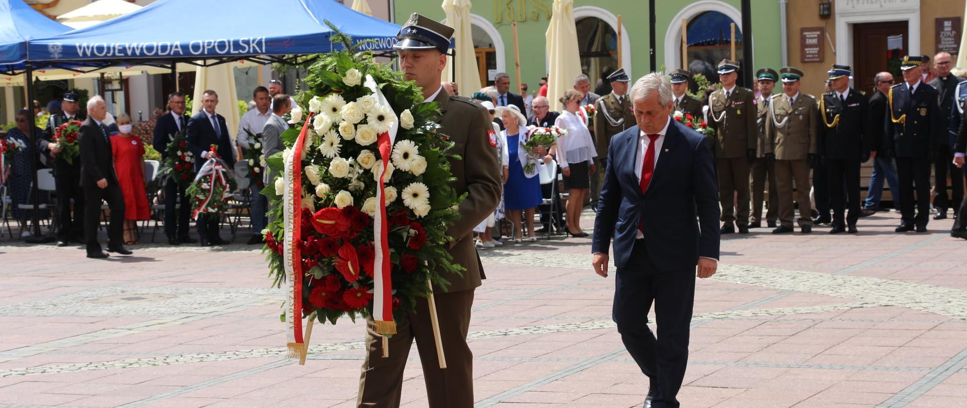 Wojewoda opolski składa wieniec w imieniu Marszałek Sejmu RP. Na zdjęciu na pierwszym planie żołnierze, który idzie w kierunku tablicy upamiętniającej wywózkę Polaków na Sybir. W dłoniach trzyma okazałą wiązanka kwiatów. Za nim wojewoda opolski, który idzie w kierunku tablicy. Spogląda w dół, ma pochylone czoło. W tle kamieniczki opolskiego rynku oraz pozostali uczestnicy uroczystości, którzy stoją w kolejce do możliwości złożenia kwiatów. Po lewej stornie kadru w tle dwa niebieskie namioty z napisem Wojewoda Opolski oraz krzesła dla gości. 