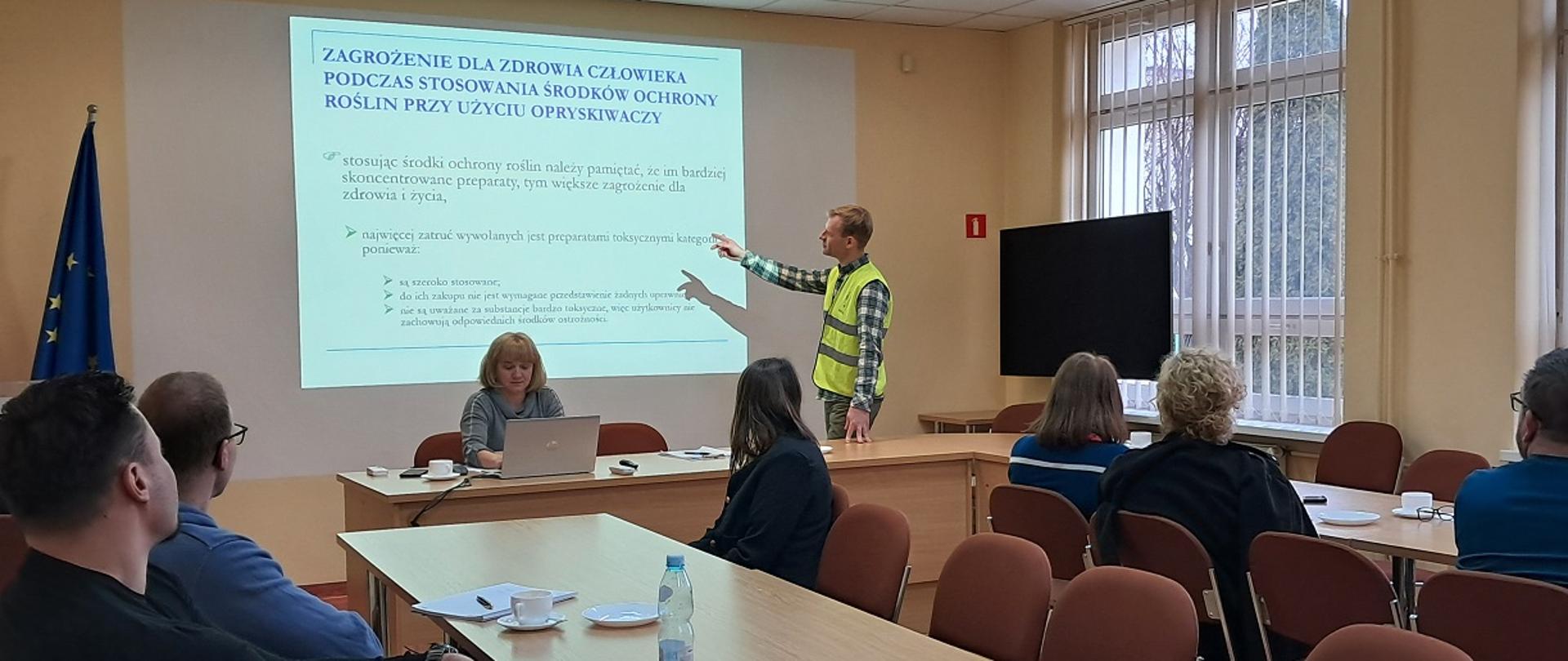 Przy ścianie stoi inspektor WIORiN i omawia tekst wyświetlony z rzutnika. Za stołem siedzi kobieta i patrzy na ekran laptopa. Za stołami siedzi sześciu uczestników szkolenia, słucha wykładowcy