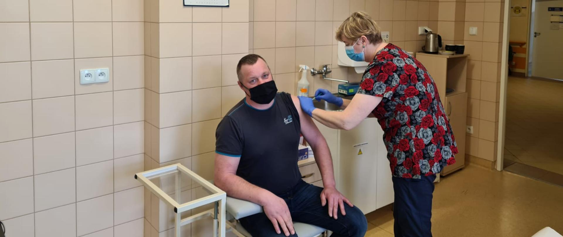 Zdjęcie przedstawia funkcjonariusza Państwowej Straży Pożarnej w pomieszczeniu szpitalnym punktu szczepień podczas wykonywanej iniekcji dawki szczepionki. Funkcjonariusz siedzi na krześle szpitalnym ubrany jeansy, koszulkę oraz maseczkę jednorazową. Pielęgniarka stojąca z boku dokonuje szczepienia.