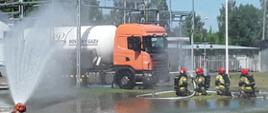 Czterech funkcjonariuszy Państwowej Straży Pożarnej w pełnym wyposażeniu spryskuje autocysternę wodą z węża strażackich. Pojazd stoi na utwardzonym placu otoczonym trawą. Dookoła terenu stoją latarnie i ogrodzenie. Po lewej, duży, strażacki zraszacz spryskuje teren wodą. Wylana woda tworzy duże kałuże dookoła cysterny.