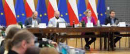 Zdjęcie sytuacyjne, szeroki kadr. Widoczny stół prezydialny, przy którym siedzą, od lewej: Współprzewodniczący Rady Michał Braun, Współprzewodniczący Rady Maciej Kunysz, Sekretarz Stanu Adriana Porowska, Dyrektor Departamentu Społeczeństwa Obywatelskiego Łukasz Jachimowicz. Na pierwszym planie widoczne rozmyte sylwetki uczestników spotkania. W tle widoczne flagi Polski i Unii Europejskiej ustawione w rzędzie za stołem.