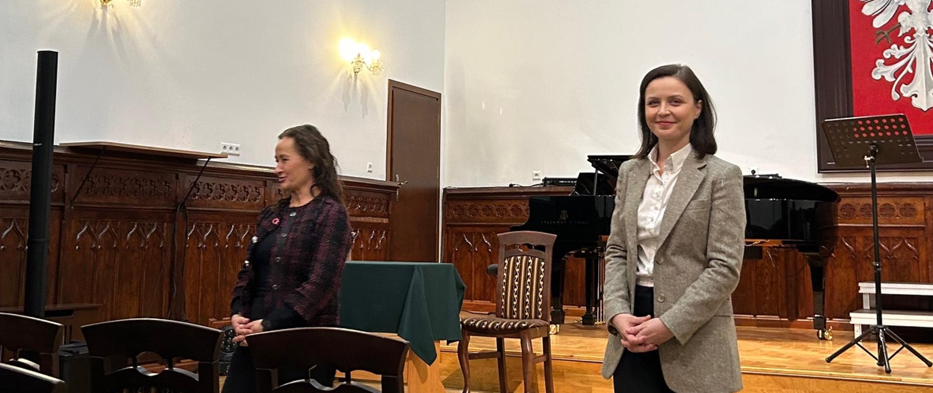 Prowadzące warsztaty wokalne stoją w sali koncertowej szkoły przed sceną z fortepianem, rozmawiając z uczestnikami siedzącymi na widowni.