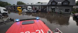 Zdjęcie wykonane z dachu czerwonego samochodu gaśniczego. Widoczny dach kabiny, na którym naniesione są numery operacyjne samochodu - 639M31. Na drugim planie zalany plac na terenie firmy. Widoczni stojący w wodzie czterej strażacy w białych hełmach, pompa szlamowa i rozwinięte węże pożarnicze.