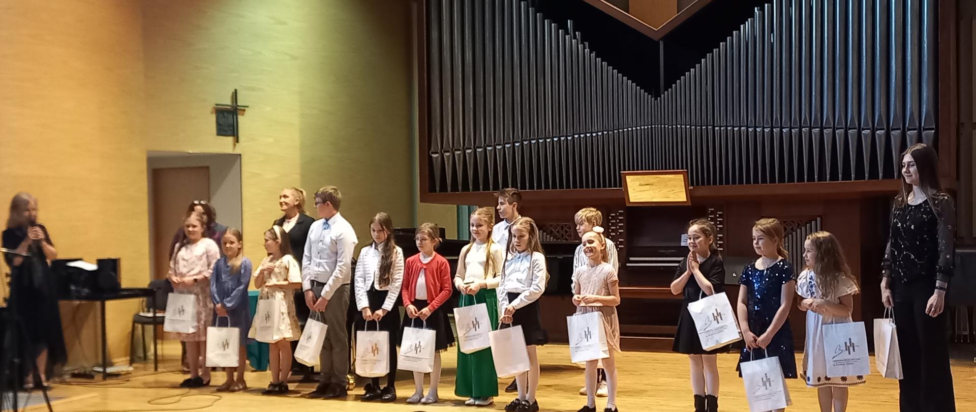 Uczestnicy konkursu na scenie sali koncertowej stoją w jednym rzędzie trzymając w dłoniach w papierowej torebce konkursowe upominki. Na pierwszym planie widownia. Za uczestnikami w tle organy oraz fortepian. Z lewej strony prowadząca konkurs oraz stolik z nagłośnieniem oraz krzesło