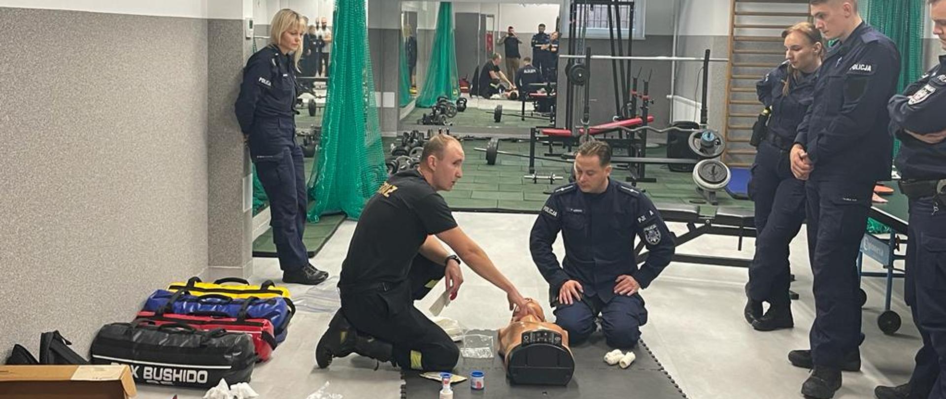 Salka gimnastyczna, siłownia. Na matach rozłożone manekiny. Instruktor strażak prowadzi szkolenie, przy nim funkcjonariusze policji