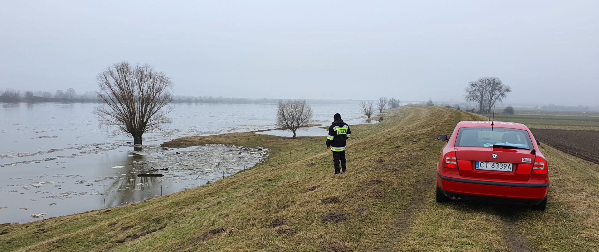 Na zdjęciu widoczny od tyłu czerwony, osobowy samochód strażacki, stojący na porośniętym trawą wale przeciwpowodziowym. W pobliżu samochodu stoi strażak ubrany na czarno z elementami odblaskowymi. Po lewej stronie płynie rzeka Wisła, z której woda wylała się na przybrzeżne łąki i podchodzi do podstawy wału. W tle widoczne drzewa, a po prawej stronie pola uprawne.
