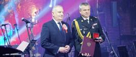 Wojewoda pomorski oraz pomorski komendant wojewódzki Państwowej Straży Pożarnej stoją obok siebie strażak prezentuje otrzymane wyróżnienie.