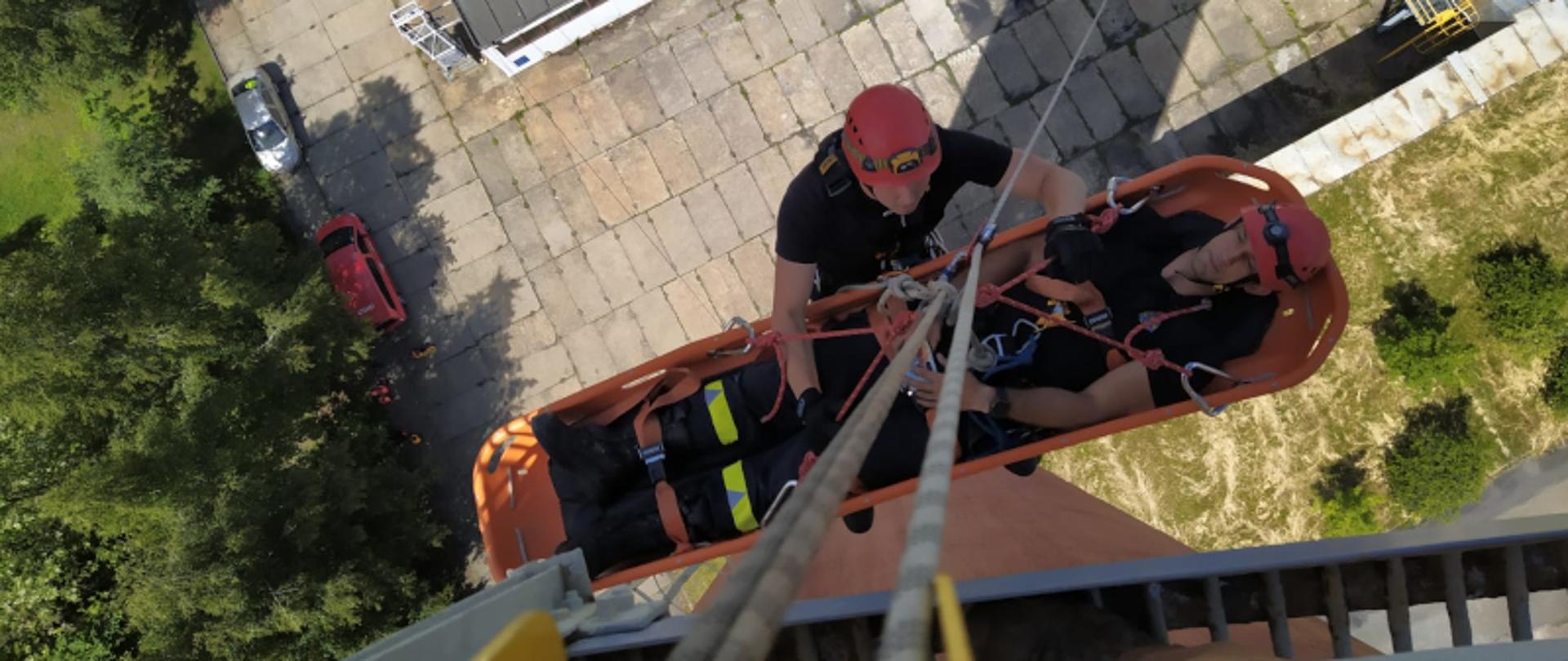 Zdjęcie robione z góry, widać dwóch strażaków podczas ćwiczeń, ubranych w czarne ubrania i czerwone kaski, jeden leży w noszach ratowniczych, drugi go asekuruje, zarówno ratownik jak i strażak leżący na noszach zawieszeni są na linach