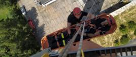 Zdjęcie robione z góry, widać dwóch strażaków podczas ćwiczeń, ubranych w czarne ubrania i czerwone kaski, jeden leży w noszach ratowniczych, drugi go asekuruje, zarówno ratownik jak i strażak leżący na noszach zawieszeni są na linach