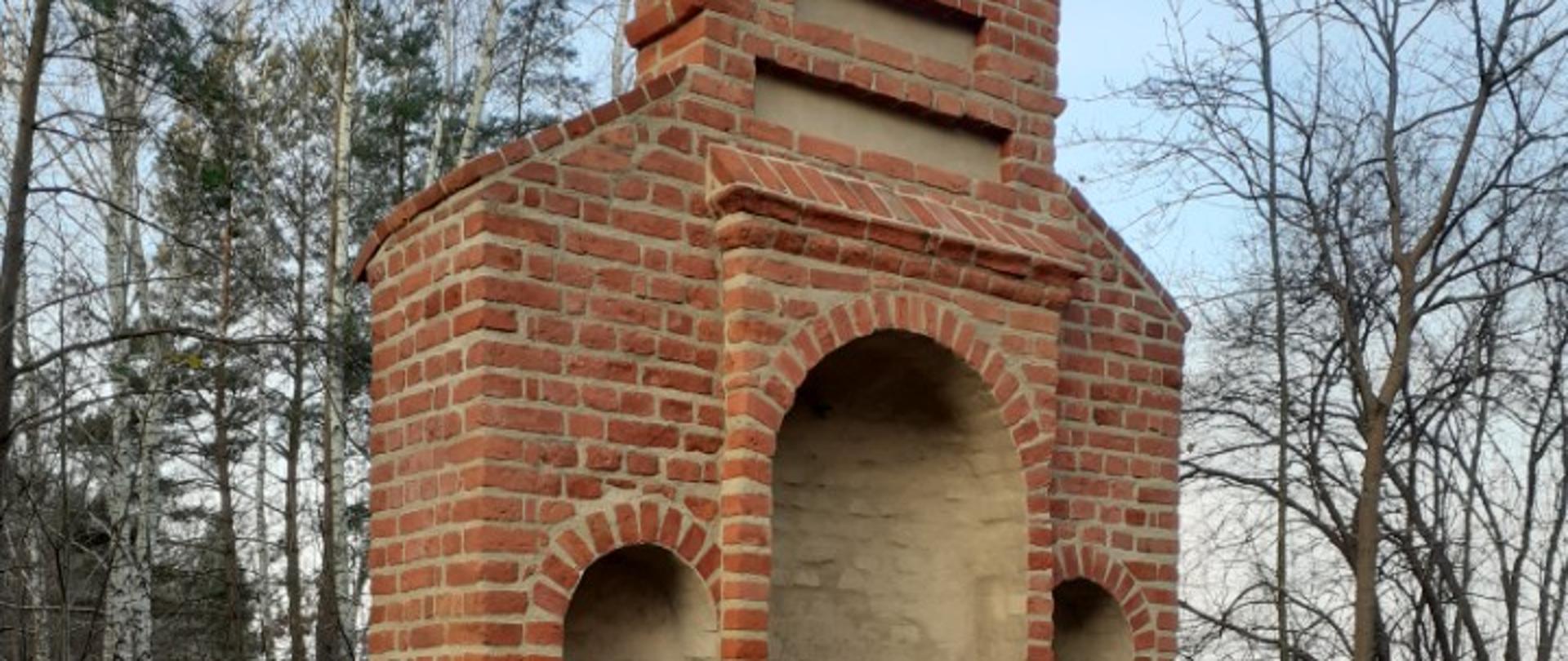 Ceglany obiekt sakralny w formie kapliczki przydrożnej, z trzema wnękami o łukowych zakończeniach, stojący na podłożu z drobnego kamienia. Kapliczka ma prostą, barokową formę, zwieńczoną górną częścią z metalowym krzyżem i ozdobnym elementem. W tle widoczne są drzewa bez liści oraz fragment ogrodzenia.