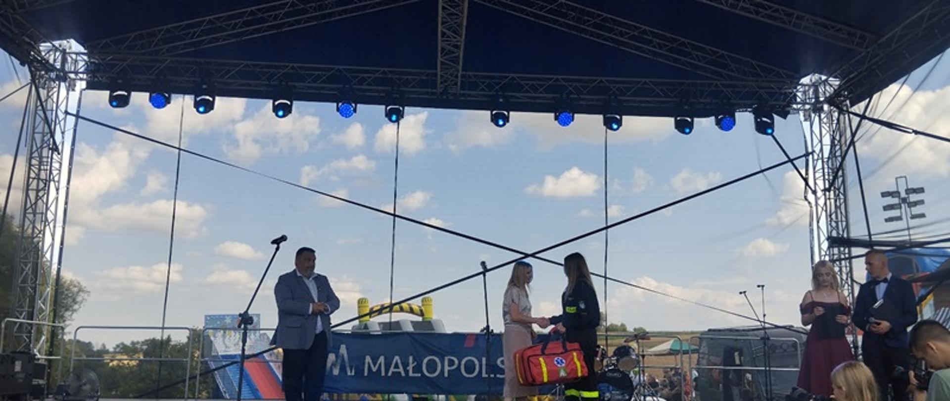 Scena plenerowa z widownią, niebem i chmurami w tle. Na scenie stoją trzy osoby – mężczyzna w garniturze z mikrofonem, kobieta w jasnej sukience oraz kobieta w ciemnym stroju trzymająca czerwoną torbę ratowniczą. Na froncie sceny widoczny jest baner z napisem „MAŁOPOLSKA”. Widać oświetlenie sceniczne i metalową konstrukcję z głośnikami