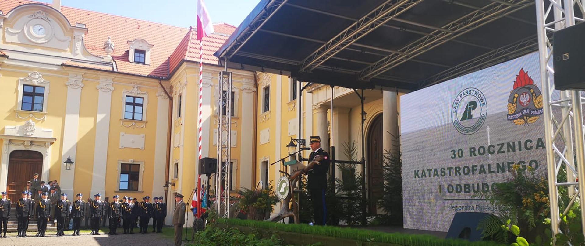 Mężczyzna w mundurze generalskim przemawiający na uroczystości. W oddali budynek z żółtą elewacja oraz stojące przed nim pododdziały uczestniczące w uroczystości