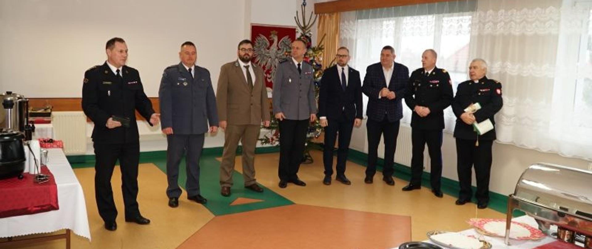 Zdjęcie przedstawia zebranych na spotkaniu wigilijnym gości. Na środku stoją Komendanci i przedstawiciele służb biorących udział w spotkaniu. W tle choinka i godło Polski.