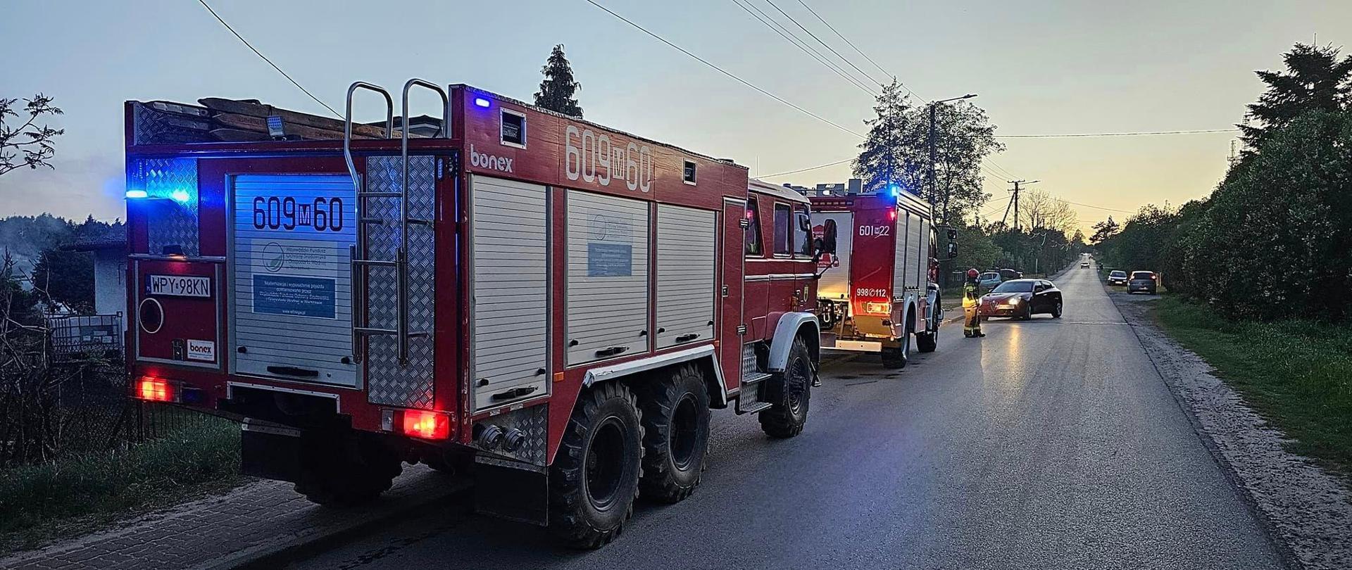 Duży, czerwony wóz strażacki z włączonymi niebieskimi światłami awaryjnymi stoi na drodze. Obok niego, nieco dalej, widać drugi pojazd, a w oddali inne samochody. Scena rozgrywa się o zmierzchu, z ciemniejącym niebem w tle. Za nimi widać strażaka w piaskowym ubraniu i czerwonym hełmie.