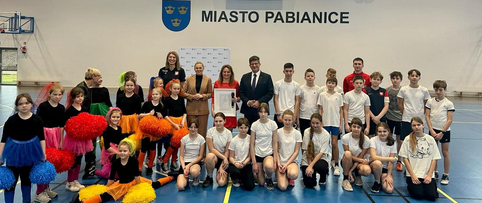 Minister Barbara Nowacka pozuje do zdjęcia z grupą dzieci na sali gimnastycznej 