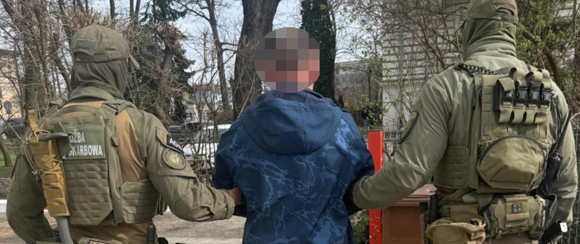 Dwóch funkcjonariuszy w umundurowaniu eskortuje osobę z zasłoniętą twarzą na terenie zewnętrznym, wśród drzew i zabudowy.