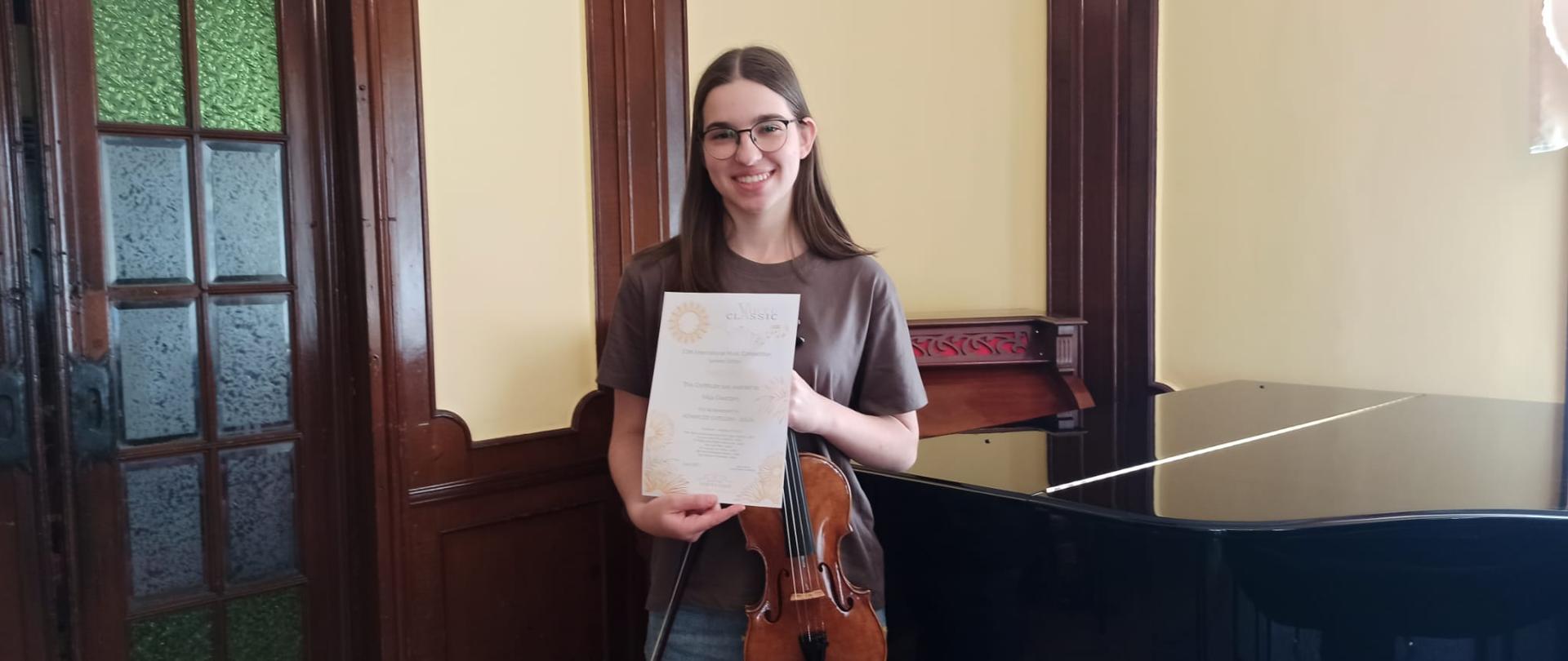 Zdjęcie kolorowe. Ciemnowłosa dziewczyna ze skrzypcami trzyma w rękach dyplom. Po prawej stronie fortepian, po lewej zabytkowe drzwi ze szprosami.