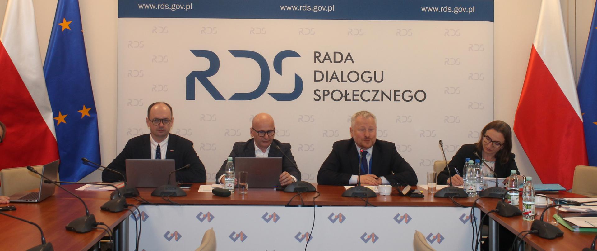 Na zdjęciu widnieje stół prezydialny za którym siedzą od lewej strony: Sebastian Gajewski - Podsekretarz Stanu w Ministerstwie Rodziny, Pracy i Polityki Społecznej, Norbert Kusiak w zastępstwie za Przewodniczącego zespołu ds. budżetu, wynagrodzeń i świadczeń socjalnych – Leszka Miętka, Jurand Drop – Podsekretarz Stanu w Ministerstwie Finansów oraz Joanna Stachura – Dyrektor w Ministerstwie Finansów i Gospodarki. Po obu stronach stołu widnieją dwie flagi: Polski i UE. W tle siedzących znajduje się baner z napisem Rada Dialogu Społecznego a nad nim wisi godło Polski.