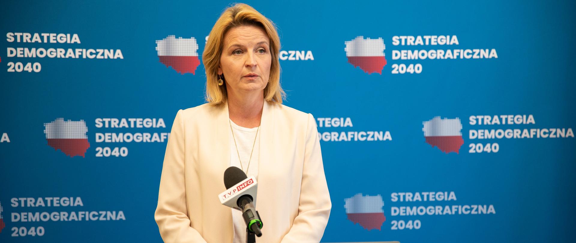 Postać minister Sochy na tle baneru z logiem w kształcie Polski wypełnionym biało-czerwonym kolorem. Obok napis Strategia Demograficzna 2040. Minister stoi przy mównicy na której widnieje herb Polski