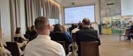 grupa osób siedzi w rzędach krzeseł, zwróceni są do stojącego w głębi zdjęcia mężczyzny, który za plecami ma wyświetloną prezentację