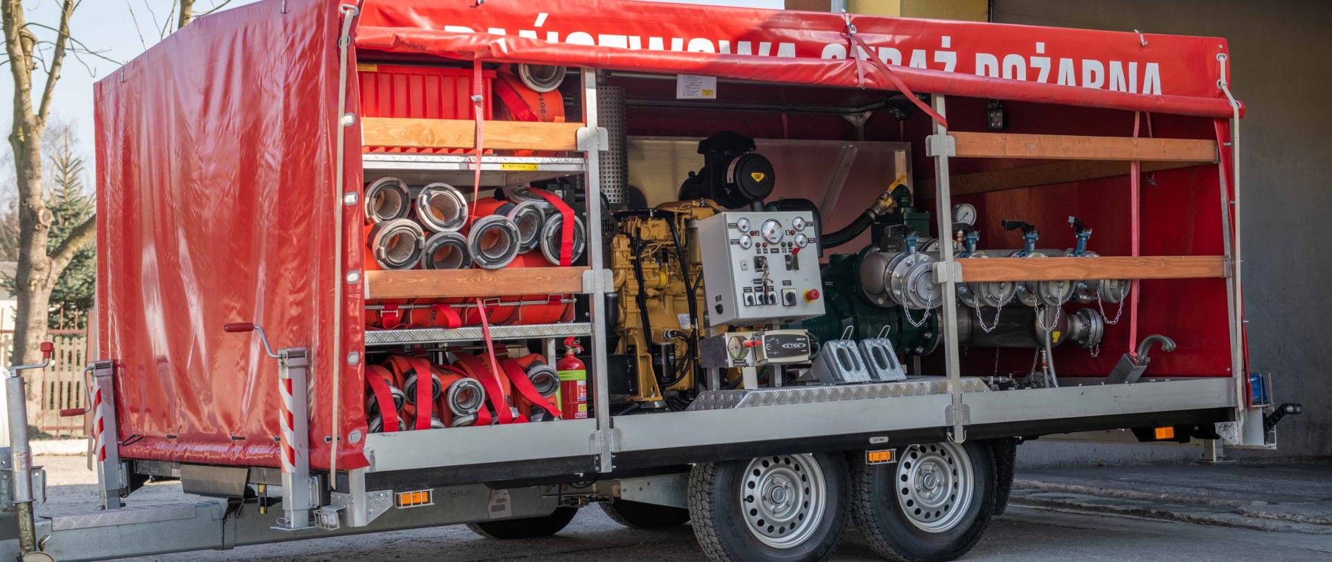 Widoczna jest pompa z czterema nasadami tłocznymi i węże tłoczne zwinięte w kręgi. Wszystko umieszczone jest na przyczepie dwuosiowej typu tandem z czerwoną plandeką znajdującej się na placu wewnętrznym przed garażem.