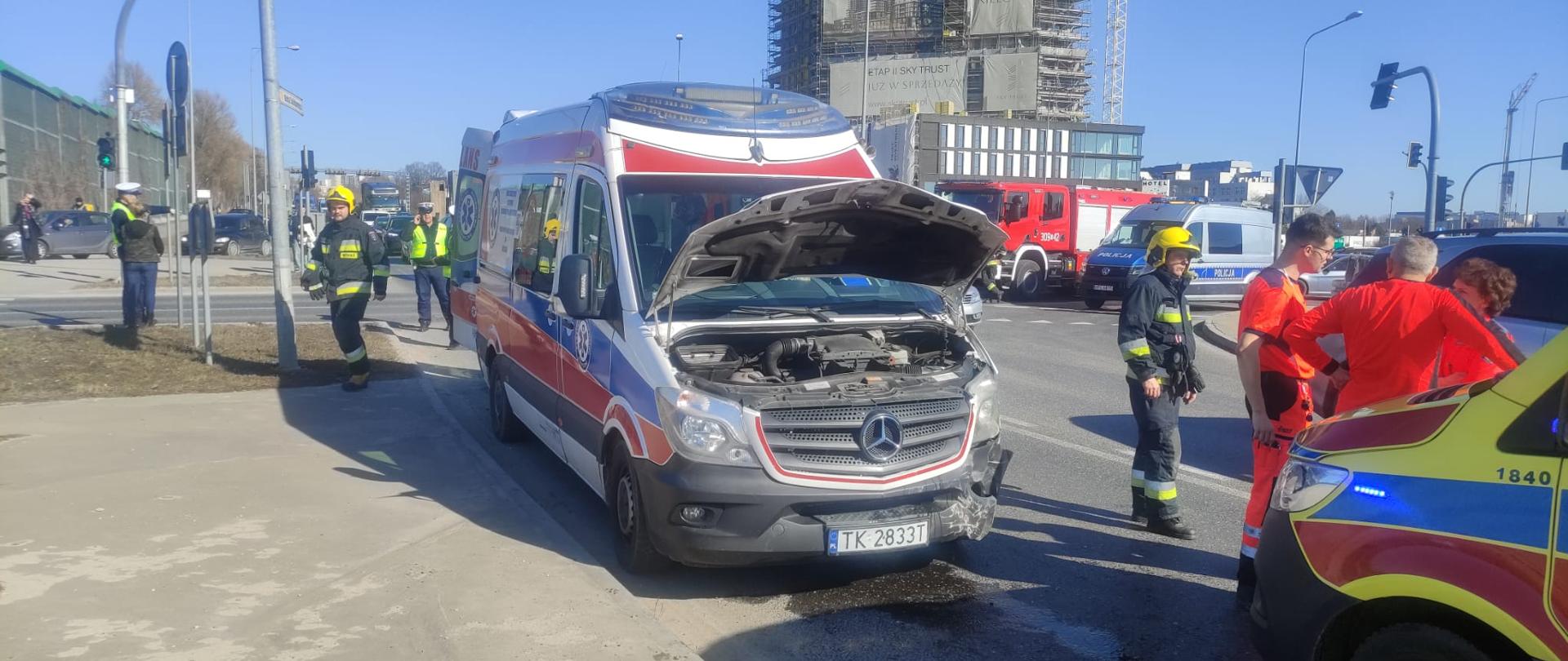 Zdjęcie przedstawia miejsce wypadku: komunikacyjnego: w centrum uszkodzona karetka, a z prawej strony samochody służb ratowniczych.