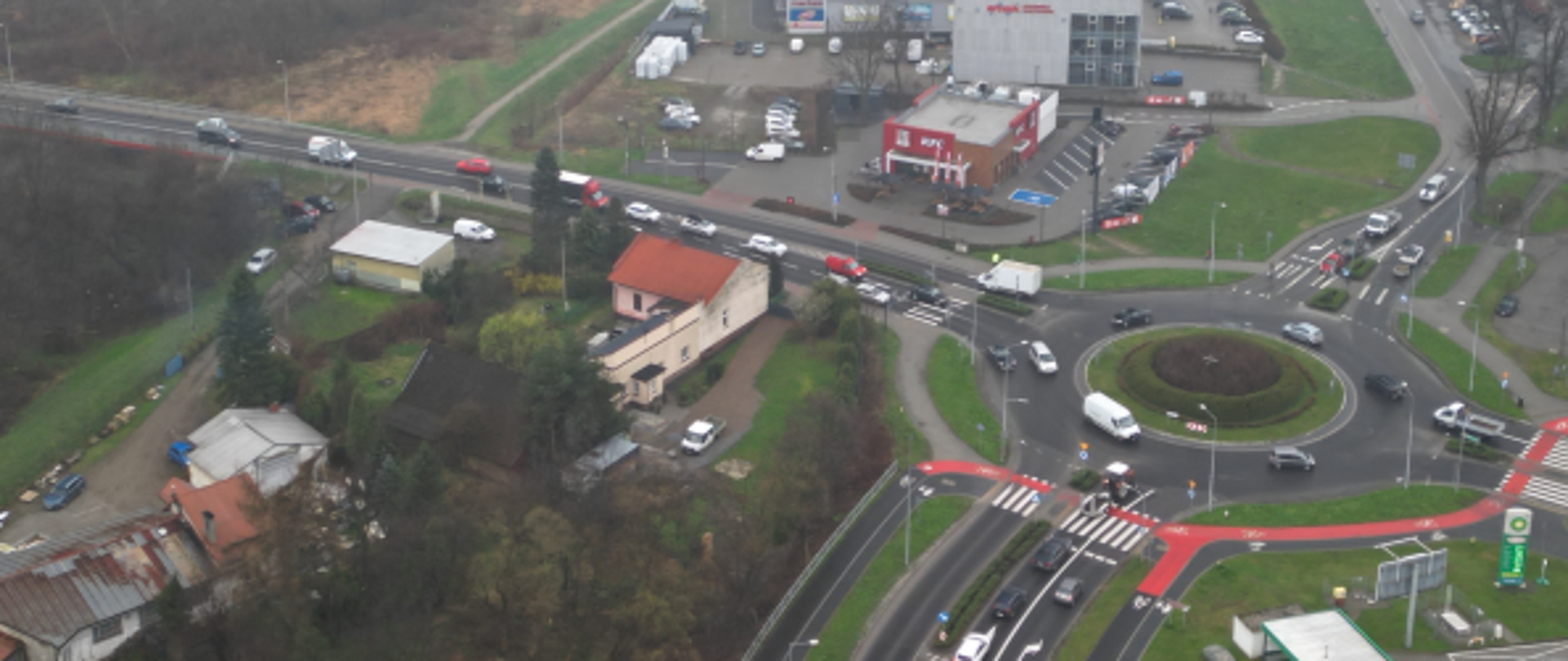Ujęcie z lotu ptaka przedstawiające skrzyżowanie dróg z rondem w małym mieście, z widocznym ruchem samochodowym, pobliskim centrum handlowym z parkingiem oraz zabudową jednorodzinną; w tle widoczna rzeka i tereny zielone spowite lekką mgłą.