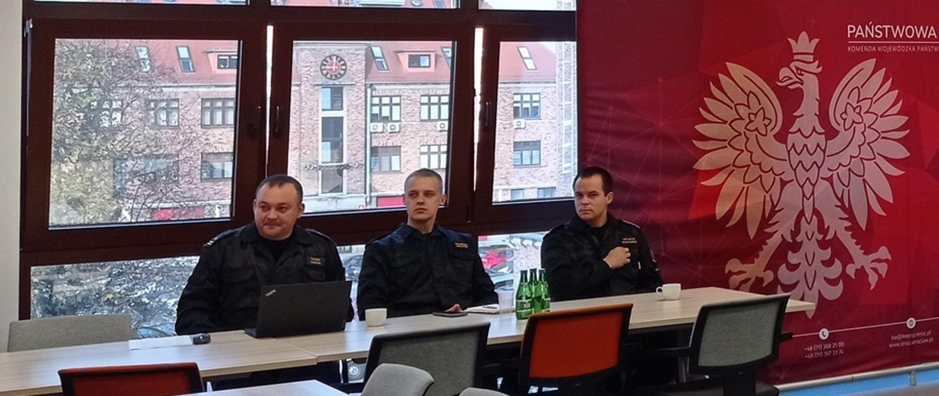 grupa strażaków w czarnych mundurach siedzi przy stołach, za nimi okna, z boku czerwona ścianka z orłem