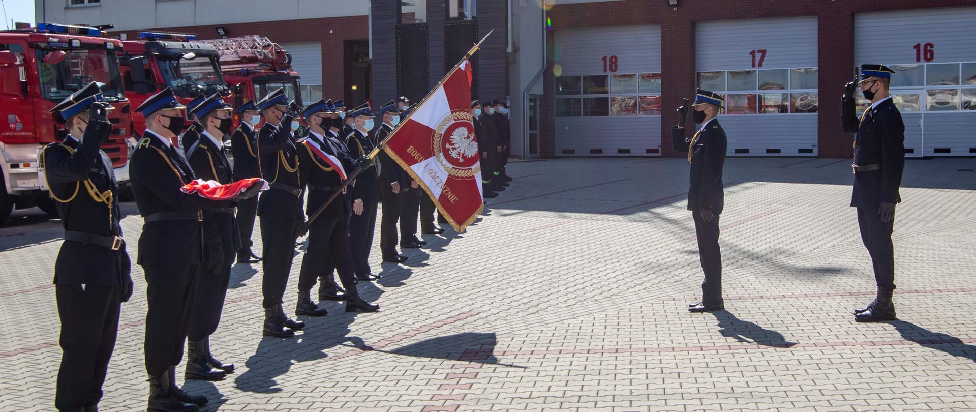 Grupa ok 20 strażaków z Państwowej Straży Pożarnej ubranych w umundurowanie wyjściowe w czapce rogatywce ze sznurem w maseczkach stoi w dwuszeregu. Pierwszy strażak od lewej salutuje, kolejny stoi z wyciągniętymi rękami na których ma złożoną flagę Państwową. Piąty od strony lewej zdjęcia strażak przepasany biało- czerwoną wstęgą stoi w „wykroku” i trzyma pochylony do przodu Sztandar Komendy Powiatowej PSP w Bochni. Naprzeciw szyku strażaków stoją dwaj strażacy ubrani w mundur wyjściowy ze sznurem, oddając honory na sztandar. Ceremonia ta odbywa się na placu wewnętrznym Komendy PSP w Bochni na tle czerwonych samochodów pożarniczych oraz budynku Komendy Powiatowej PSP w Bochni