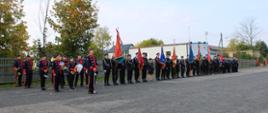 orkiestra dęta osp oraz sztandary jednostek osp stojące w szeregu w czasie uroczystości, w tle drzewa i budynki