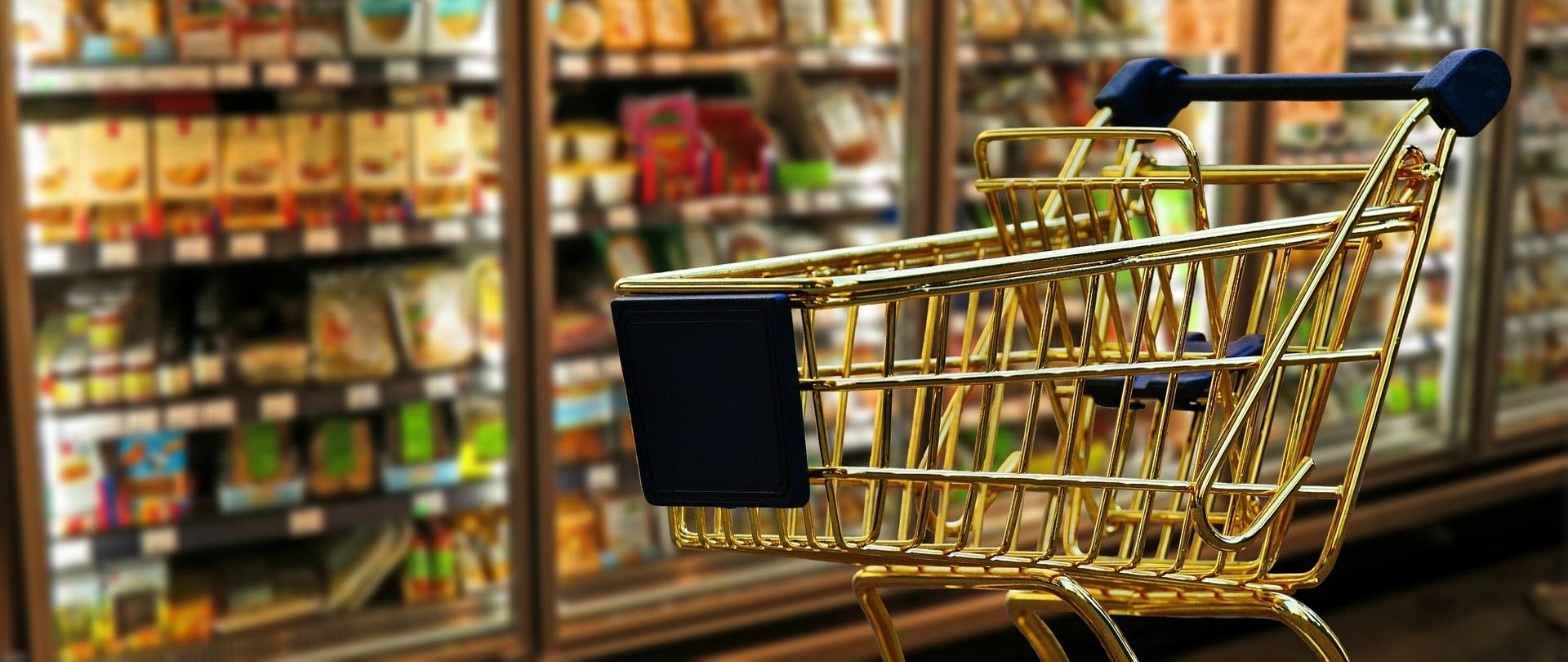Na zdjęciu jest duży wózek na zakupy, w tle lodówki z produktami spożywczymi.