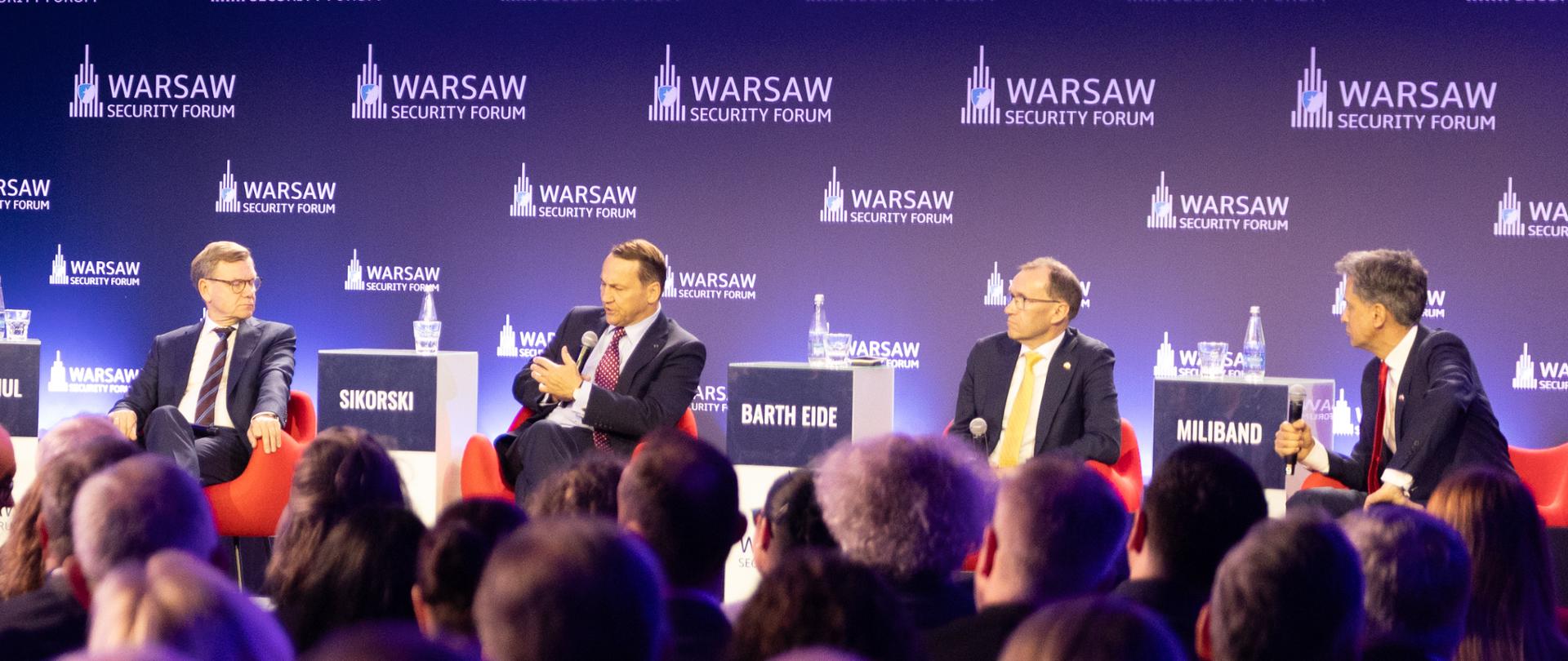 Radosław Sikorski, Deputy Prime Minister and Minister of Foreign Affairs of Poland, participates in a panel discussion at the Warsaw Security Forum. He is seated on stage alongside the other panellists, while his image is displayed on a large screen to the left. The event’s logos and purple and blue stage lighting are visible in the background.