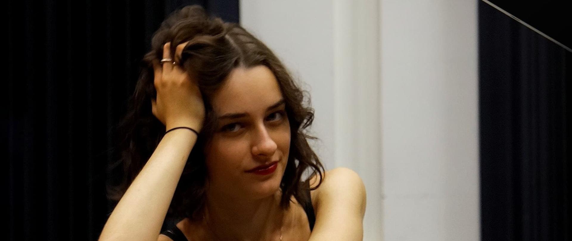 Tło obrazka czarne. U góry biały napis: "Recital fortepianowy Nikoli Zwara", dalej do dołu po prawej stronie "3 października 2024 godz. 17:00". Poniżej kobieta opierająca się na fortepianie. Na fortepianie biały napis: " Sala koncertowa Państwowej Szkoły Muzycznej I stopnia w Kościerzynie". U samego dołu biały napis: "w programie: J. S. Bach, F. Chopin, F. Liszt, S. Prokoliev".