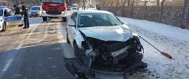 Zdjęcie przedstawia drugi uczestniczący w zdarzeniu pojazd. To małe auto osobowe koloru srebrnego. Auto ma całkowicie zniszczony przód. W tle widać radiowóz policyjny, policjantów i pojazdy, które nie mogą przejechać